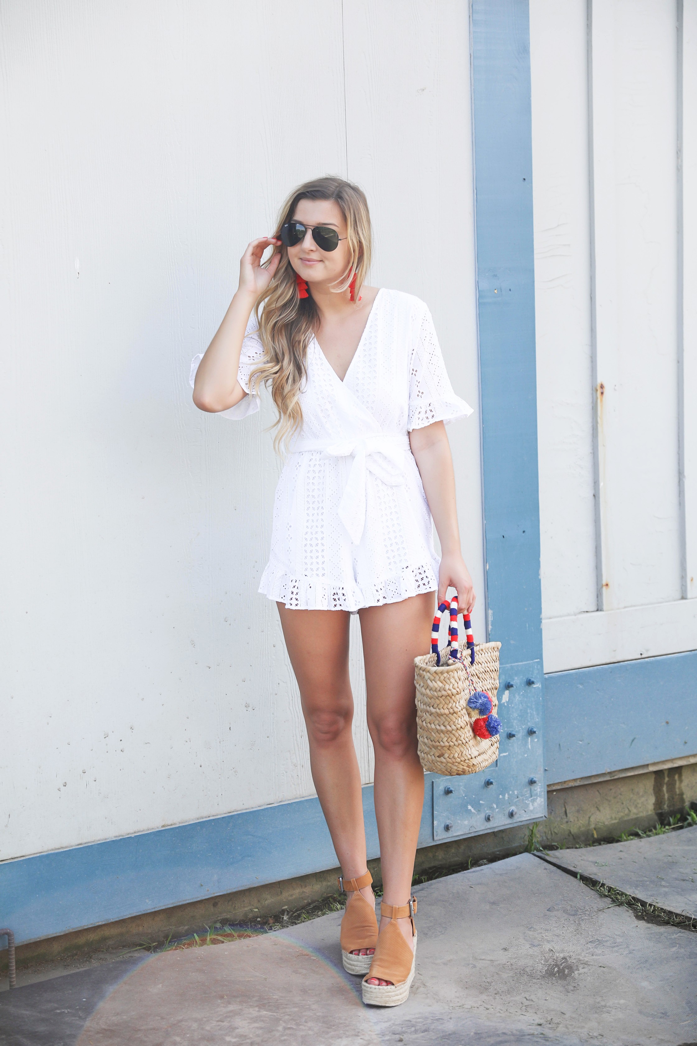 White ruffle tied romper perfect for dressing up or dressing down! Paired with a cute straw beach bag for fourth of july! The beach bag has red white and blue pom poms on it! I finished the fourth of july outfit with red tassel earrings and my favorite wedges! Details on fashion blog daily dose of charm by lauren lindmark